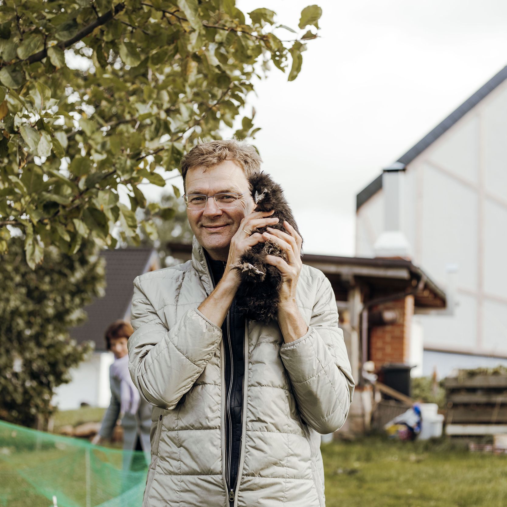 Roger Herold lächelnd auf seinem Hof mit Huhn im Arm, im Hintergrund Baum, Schuppen und Haus