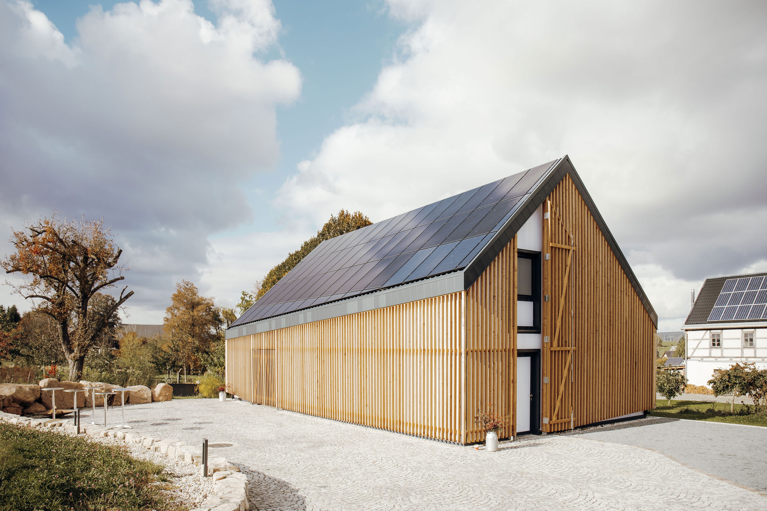 Modernes Haus mit Holzverkleidung, asymmetrischem Spitzdach und Solarpaneelen auf dem Dach. Dahinter Bäume und Himmel mit Wolken.